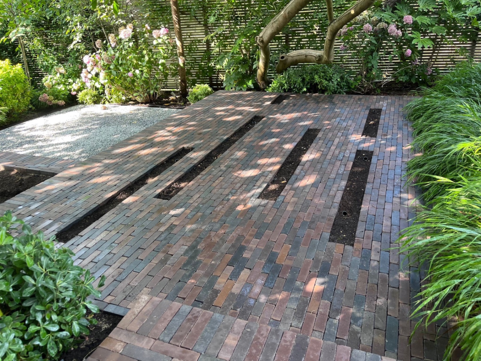 Broad clay pavior terrace with rectangular planting inserts cut into the paving and a gravel landing beyond, framed by hydrangeas and a slatted fence