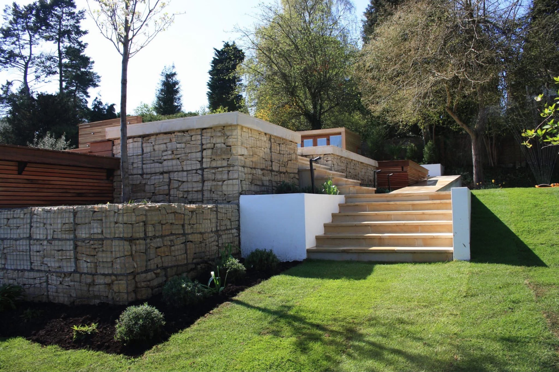 Completed hillside terracing, with gabion cage retaining walls topped with sandstone coping, broad sandstone steps up the slope and a white rendered wall