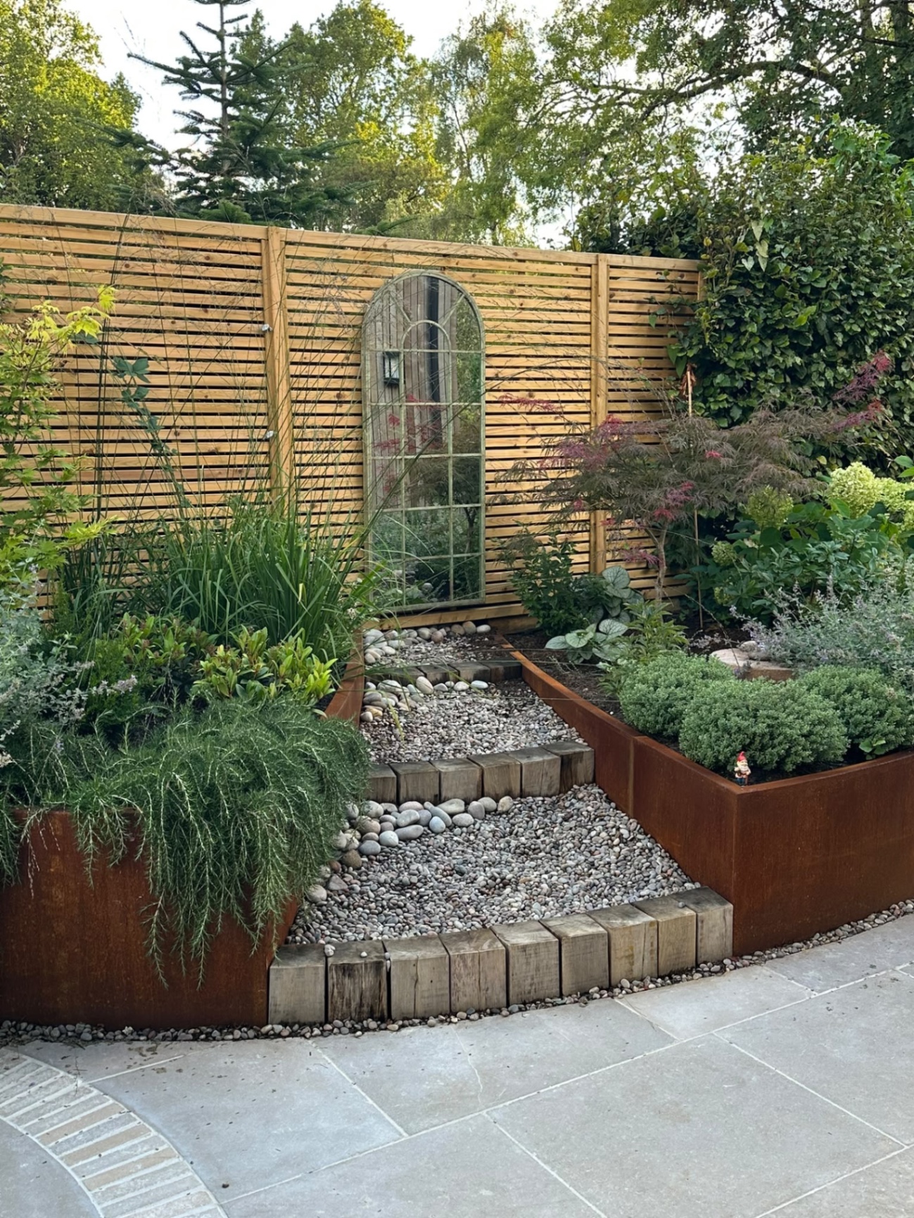 Tiered pebble path and timber-block steps rising between raised corten steel planters toward the garden mirror