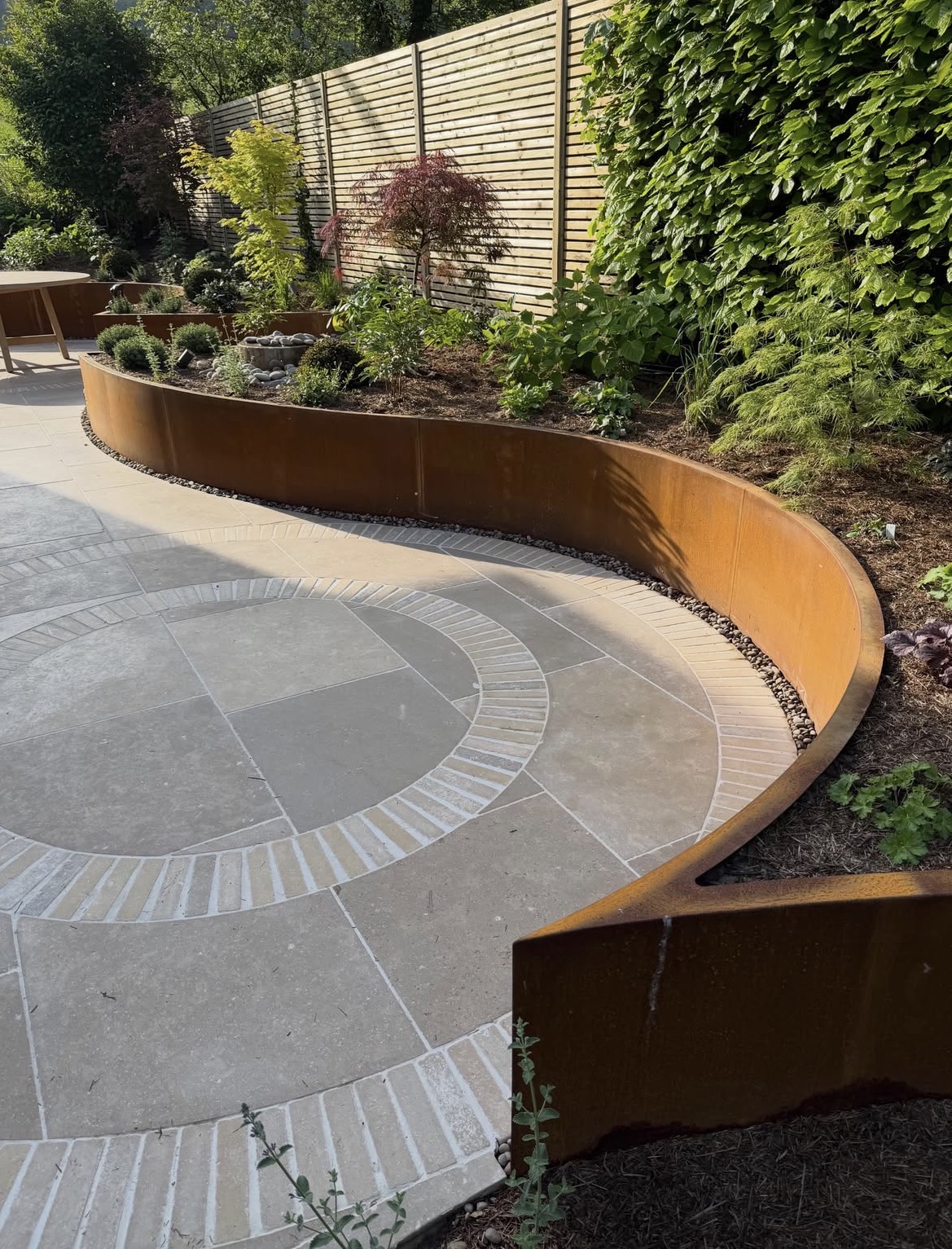 Curved corten steel planting bed sweeping around a circular paved terrace