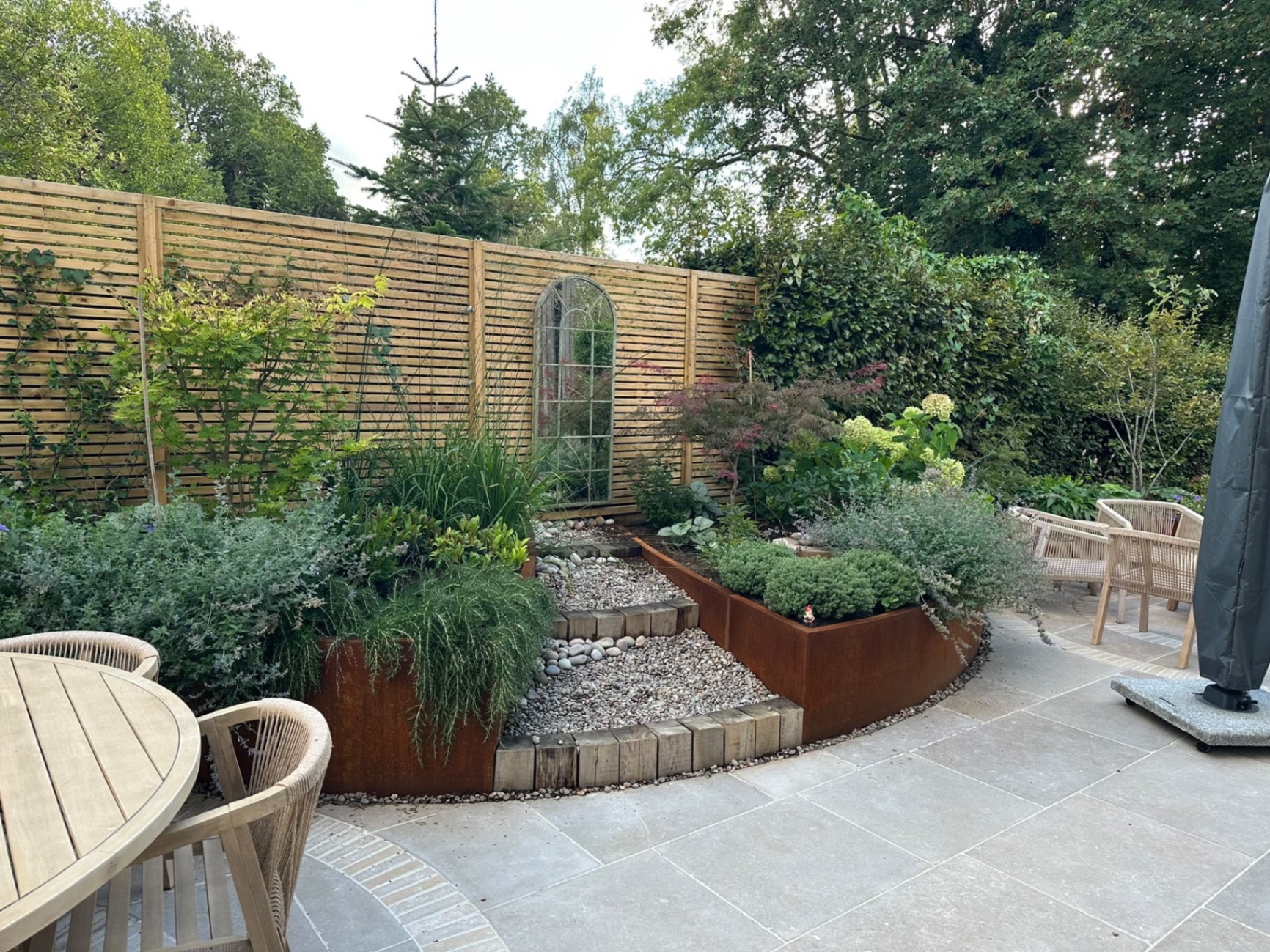 Pebble dry-stream path with timber-block steps winding between corten steel planters toward an arched mirror on the fence