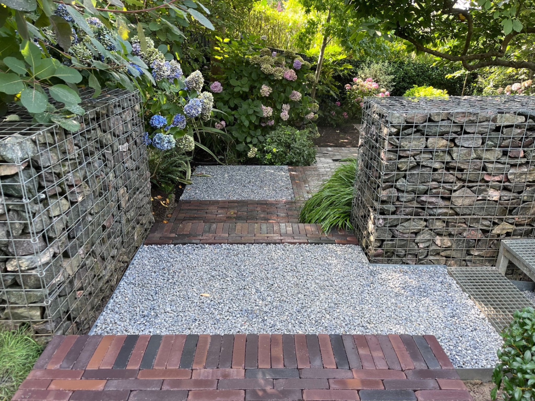 View up the sloped garden showing gabion retaining walls, rectangular gravel landings and rows of clay paviors navigating the level change