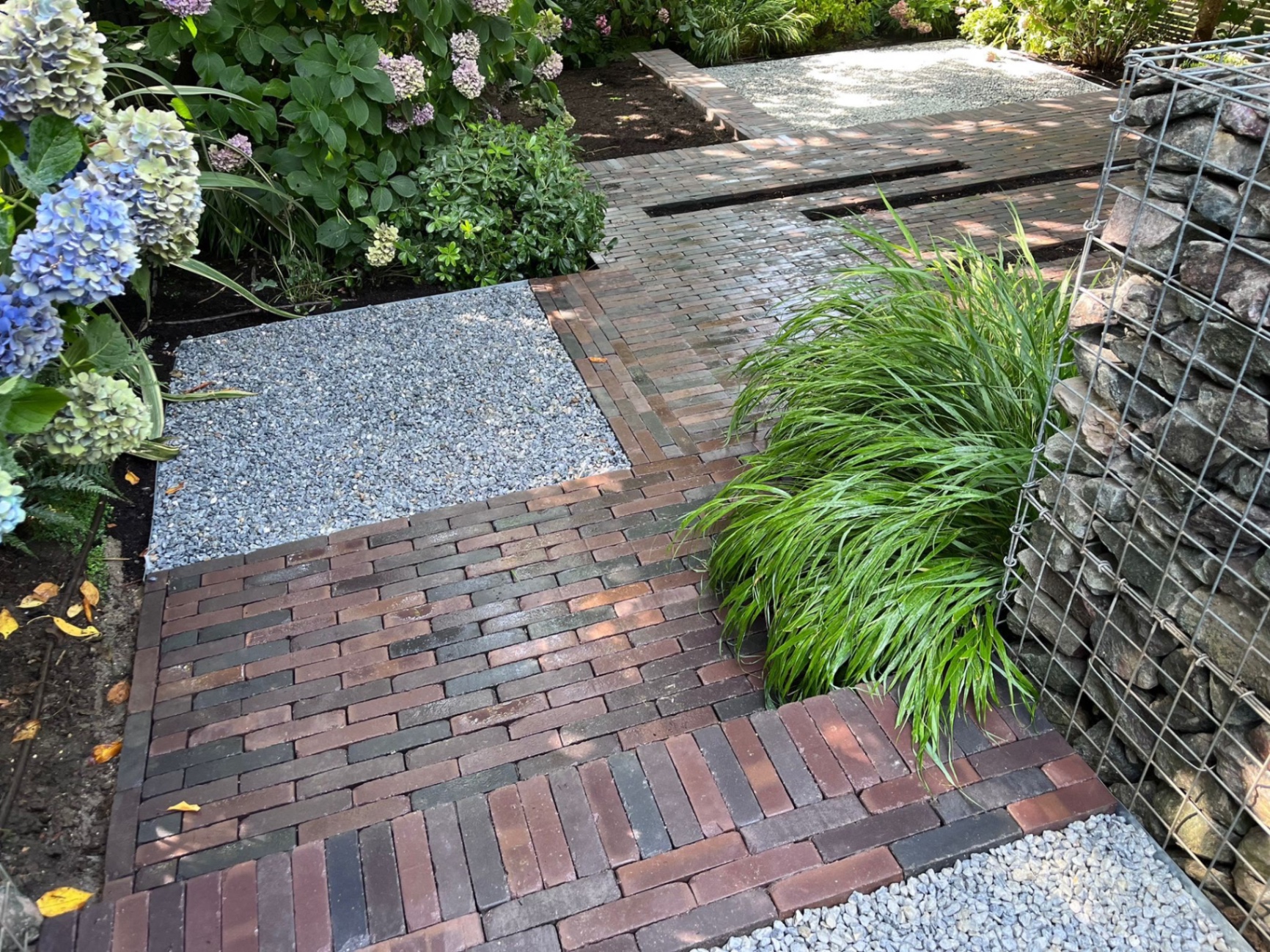 Path of clay pavior steps descending the slope through gravel landings, held by metal edging with gabion walls and hydrangeas either side