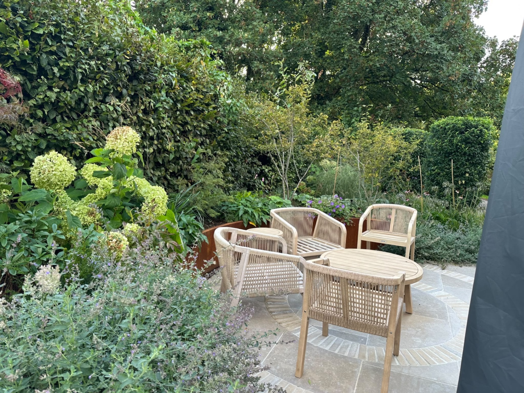 Seating area with round table on a circular brick detail, backed by a curved corten steel retaining wall and dense planting