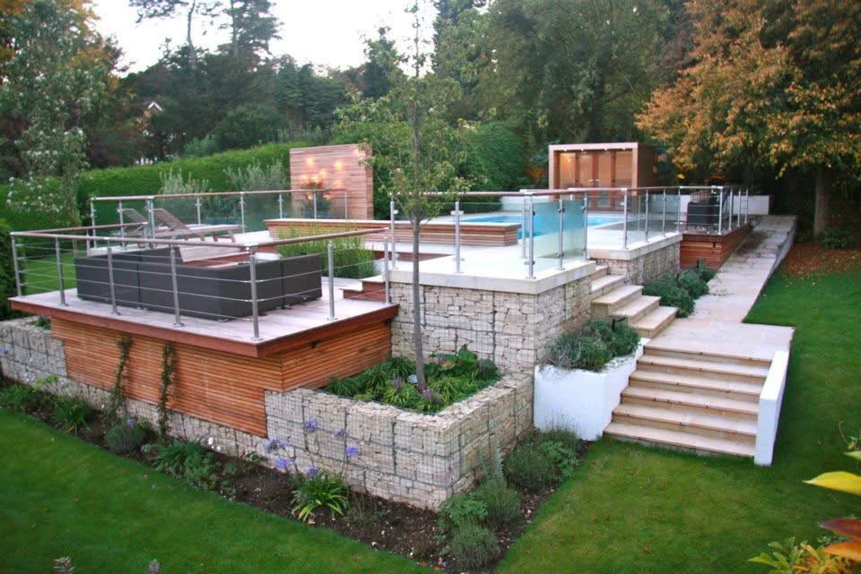Elevated daytime view across the multi-level garden, with a hardwood deck and sofa, glass balustrade, gabion retaining planters and sandstone steps around the pool